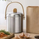 Cubo de compost de acero inoxidable junto a una bolsa de papel kraft abierta y restos vegetales para hacer compost en casa