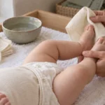 Bebé con pañal de tela sobre el cambiador mientras un adulto le sostiene los pies y una toallita de algodón