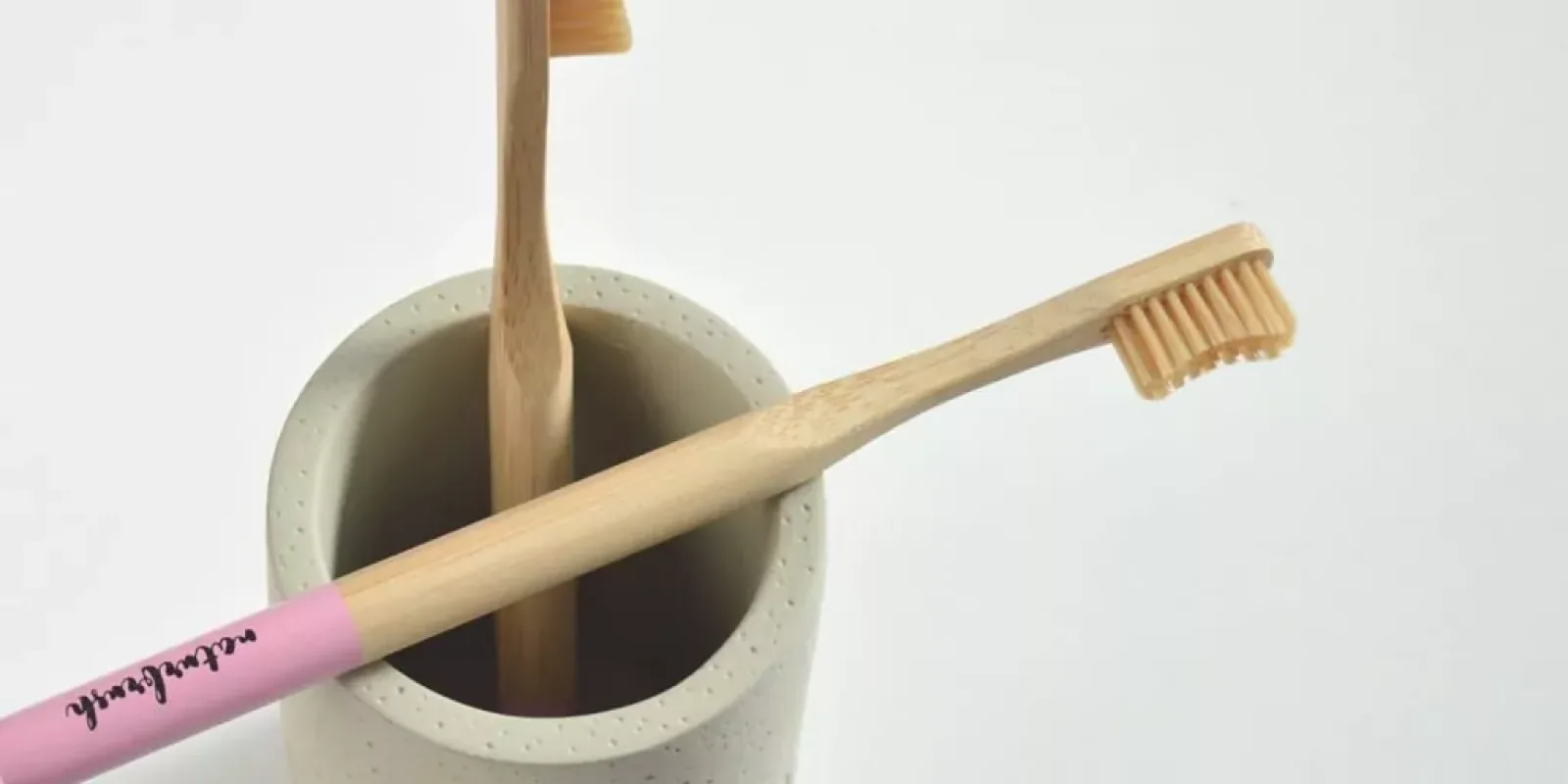 Dos cepillos de dientes de bambú, uno con mango rosa, en un vaso de cemento claro sobre fondo blanco