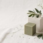 Jabón natural cúbico, ramas de olivo y flores blancas sobre fondo de lino
